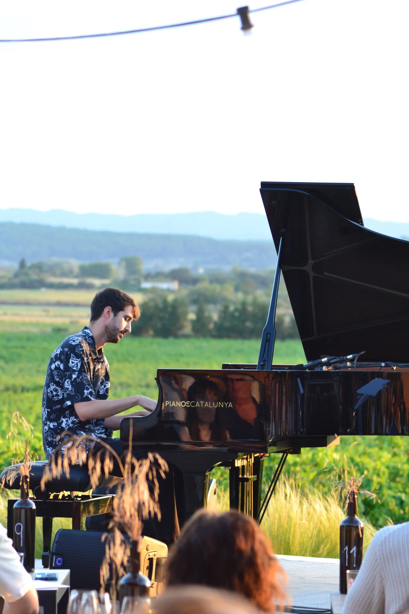 Aleix Cansell playing a grand piano outdoors, with fields and hills in the background